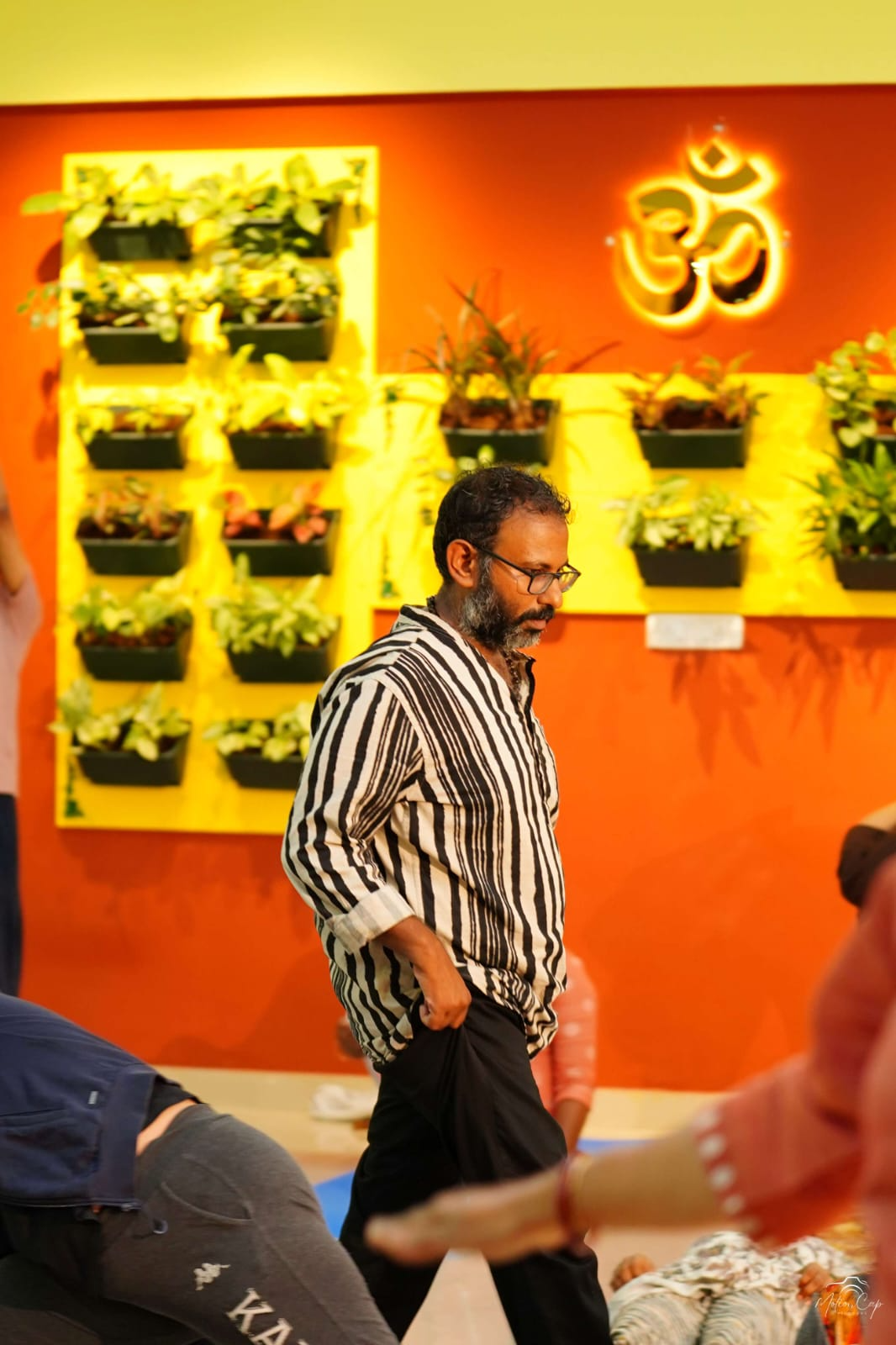 A teacher walking through the studio beneath a glowing Om symbol and wall planters