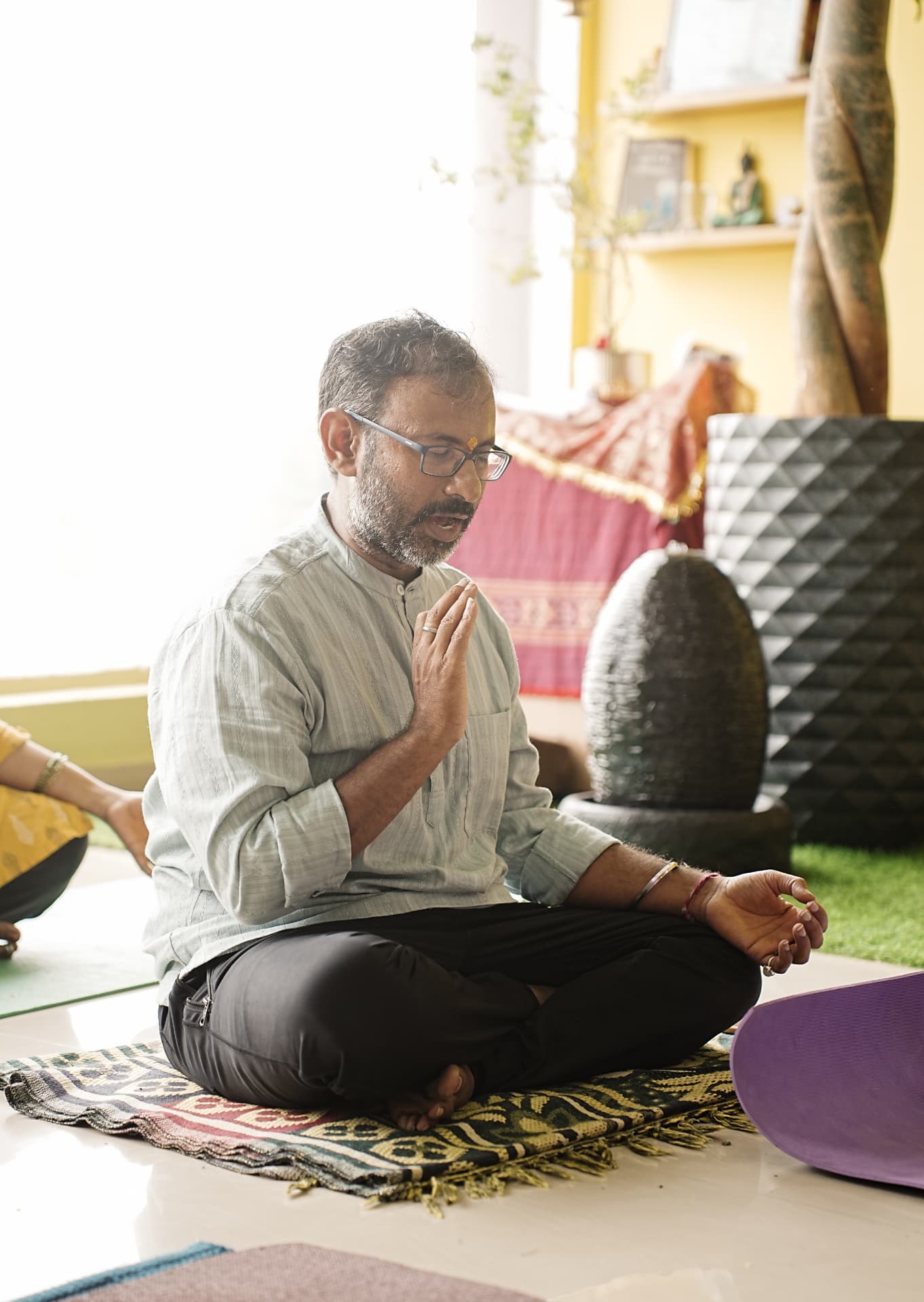 A teacher seated cross-legged chanting with hands in prayer